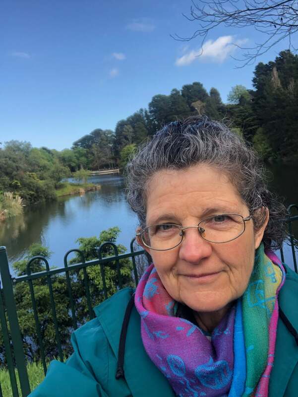 Anglo Saxon older female wearing a colorful scarf with a lake and gardens behind her - Kim Ross, Psychologist mindfully walking for health.