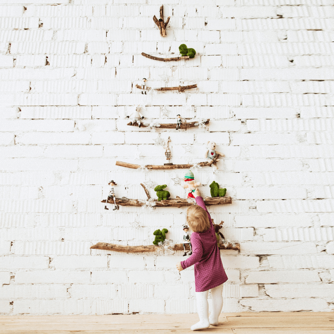 Young girl placing a chirstmas elf on a branch of a wooden tree like series of benches attached to a white wall